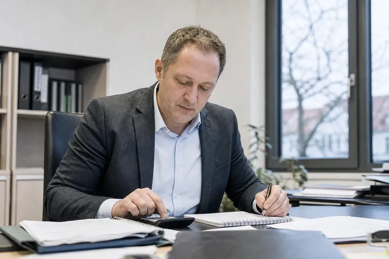 Makler kalkuliert Kosten neben einer Vorgangsmappe am Schreibtisch